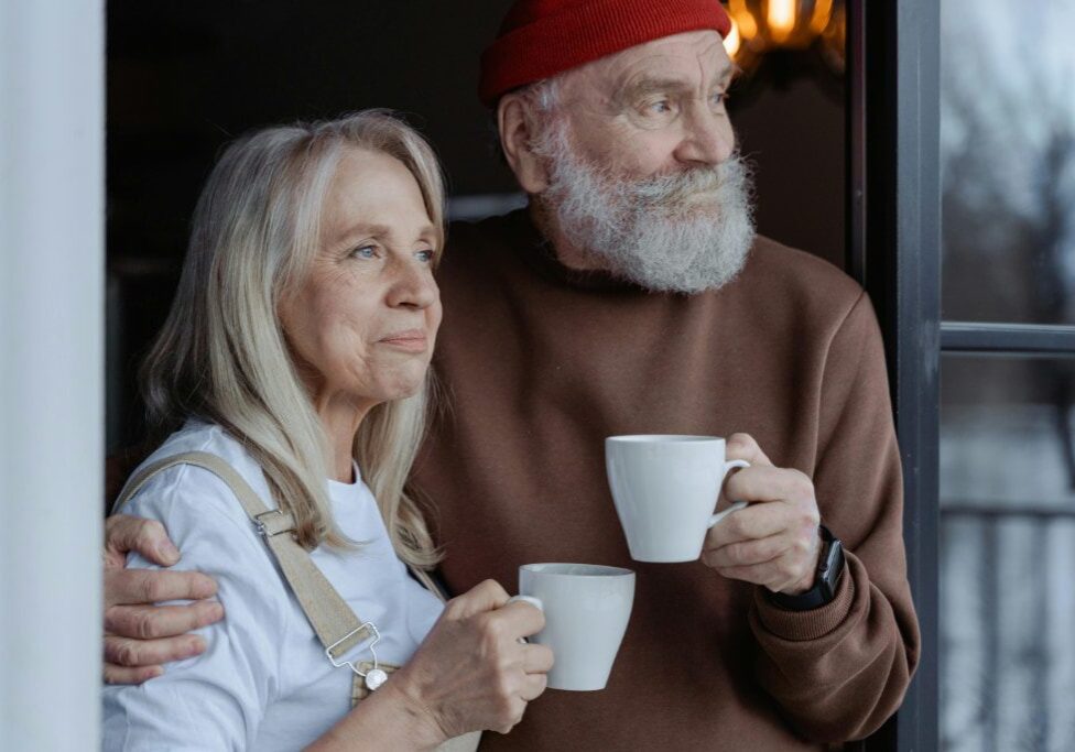 An older couple, handyman for seniors, holding mugs and looking out a window together.